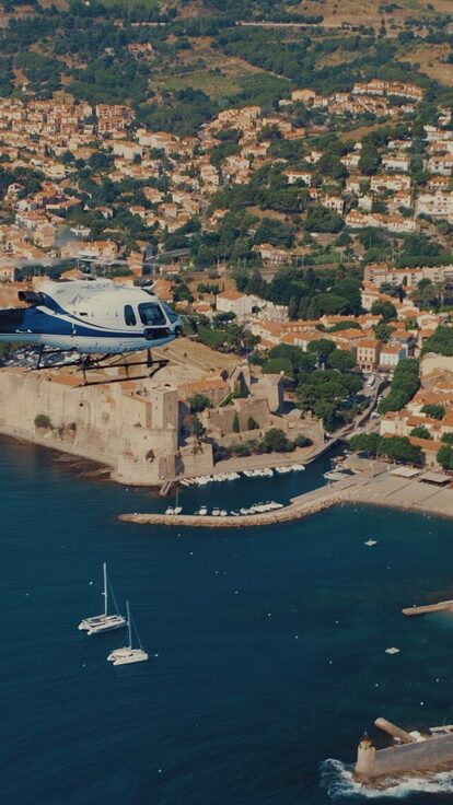 Un hélicoptère survole port, fort et collines verdoyantes.