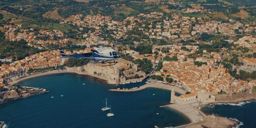 Un hélicoptère survole port, fort et collines verdoyantes.