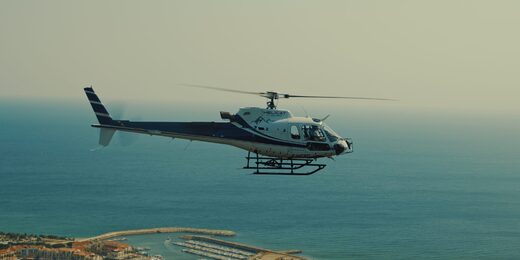 Un hélicoptère survole un port de plaisance, avec mer et côte en arrière-plan.