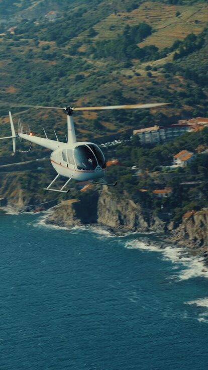 Un hélicoptère blanc survole des falaises côtières et collines verdoyantes près de l'océan.