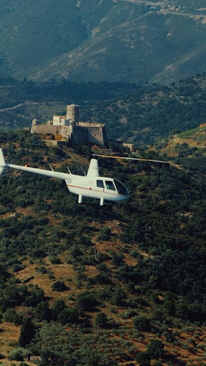 Un hélicoptère blanc survole des collines verdoyantes, avec un château derrière lui.