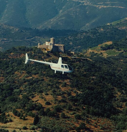 Un hélicoptère blanc survole des collines verdoyantes, avec un château derrière lui.