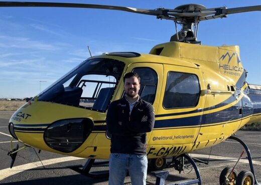 Un homme devant un hélicoptère jaune sur l'hélipad, bras croisés sous un ciel dégagé.