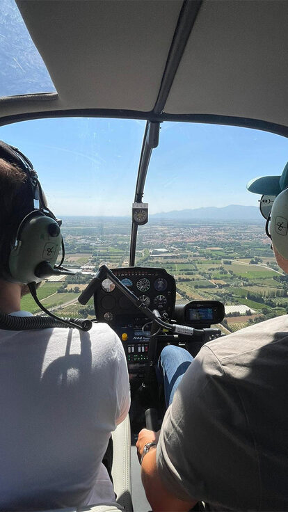 Deux personnes casquées pilotent un hélicoptère au-dessus des montagnes et champs verdoyants.