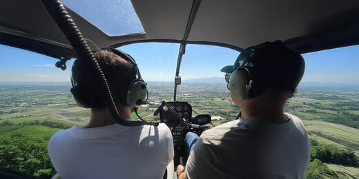 Deux personnes casquées pilotent un hélicoptère au-dessus des montagnes et champs verdoyants.